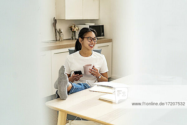 Junge Frau mit Kaffeetasse und Mobiltelefon lehnt die Beine auf den Tisch zu Hause