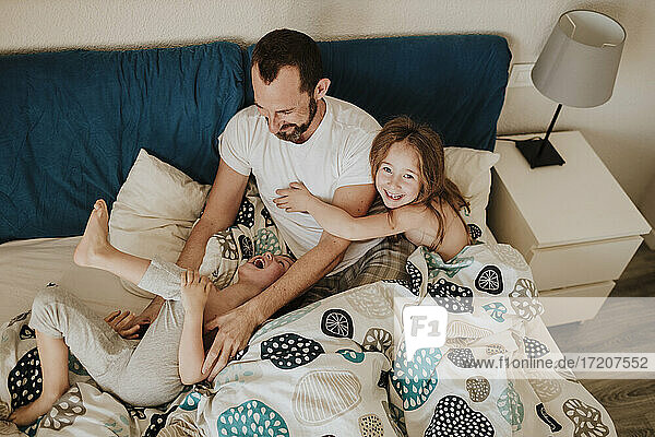 Happy father playing with cheerful son while daughter sitting on bed at home