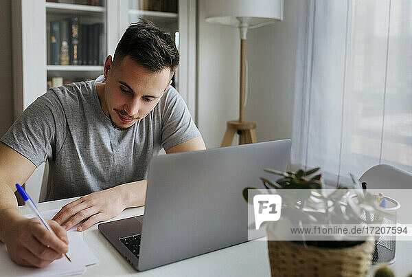 Männlicher Unternehmer  der mit einem Laptop im Büro zu Hause sitzt und auf Papier schreibt