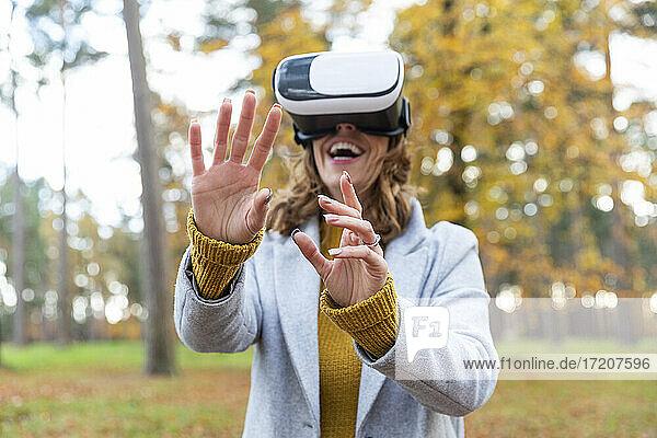 Smiling woman wearing virtual reality headset stretching hands while standing in forest