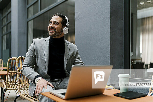 Lächelnder Geschäftsmann mit Laptop und Kopfhörern  der wegschaut  während er im Café sitzt
