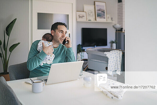 Vater mit Laptop  der seinen Sohn trägt  der wegschaut  während er am Schreibtisch im Heimbüro mit dem Handy telefoniert
