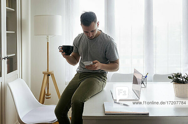 Junger männlicher Unternehmer  der einen Kaffee trinkt und dabei sein Smartphone benutzt  während er im Büro zu Hause am Schreibtisch sitzt
