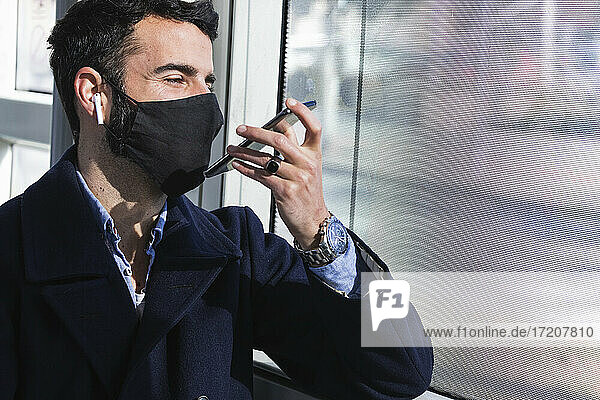 Männlicher Geschäftsmann mit Gesichtsschutzmaske  der im Zug über Lautsprecher mit seinem Smartphone spricht