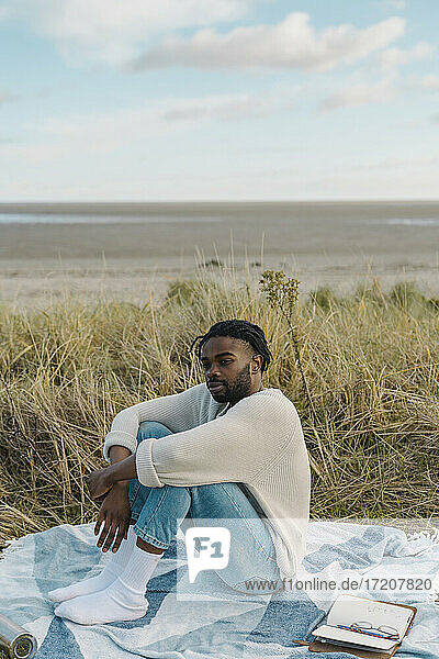 Entspannter junger Mann sitzt auf einer Decke inmitten von getrockneten Pflanzen am Strand