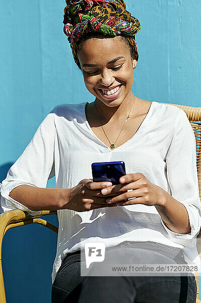 Lächelnde junge Frau mit Kopftuch  die ein Mobiltelefon benutzt  während sie auf einem Stuhl vor einer blauen Wand sitzt