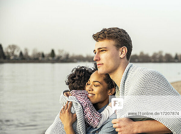 In eine Decke gehüllter junger Mann steht neben einer Frau und einem Jungen am See