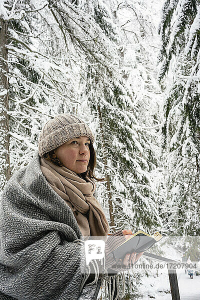 Nachdenkliche Frau in warmer Kleidung hält ein Buch  während sie im Wald steht