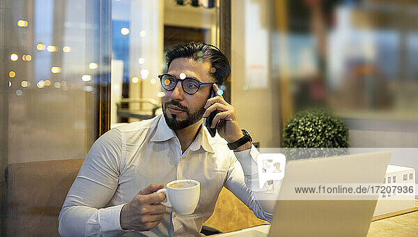 Businessman talking on smart phone while holding coffee cup in cafe
