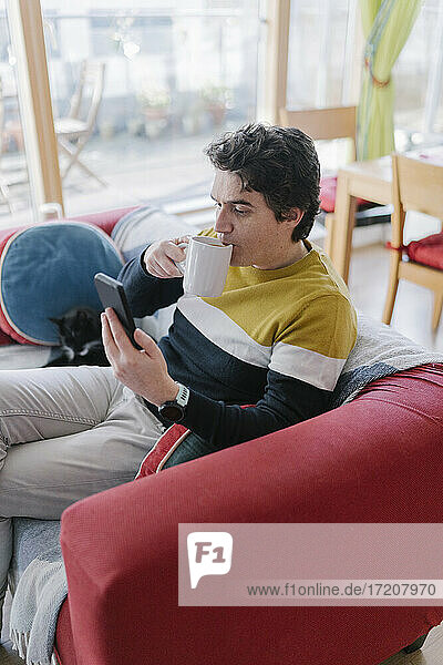 Mid adult man using mobile phone while drinking coffee while sitting on sofa at home