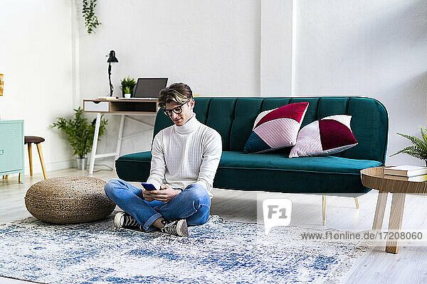 Junger Mann sitzt mit gekreuzten Beinen im Wohnzimmer und benutzt ein Smartphone