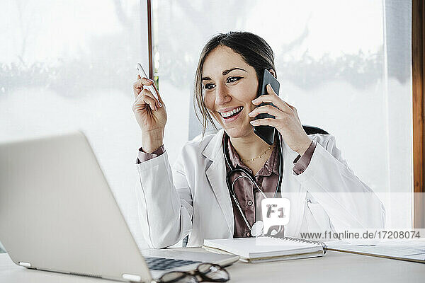 Lächelnde Ärztin  die auf einen Laptop schaut  während sie mit einem Mobiltelefon im Krankenhaus spricht