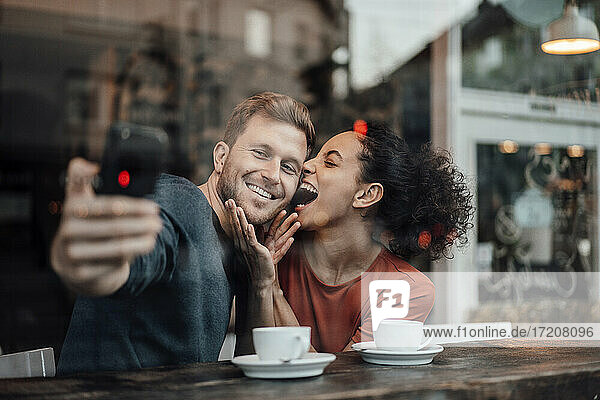 Lächelnder Freund nimmt Selfie durch Handy mit Freundin beim Sitzen im Cafe