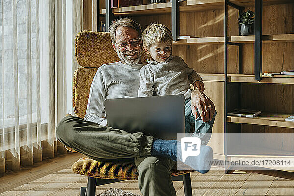 Lächelnder reifer Mann mit Sohn  der einen Laptop benutzt und auf einem Stuhl sitzt
