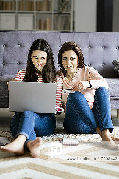 Lächelnde Tochter  die Rechnungen online bezahlt  während sie neben ihrer Mutter sitzt  die eine Kreditkarte zu Hause hält