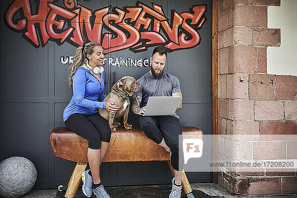 Männlicher und weiblicher Sportler  der einen Laptop benutzt und mit einer Bulldogge auf einer Bank vor einer Graffiti-Tür sitzt