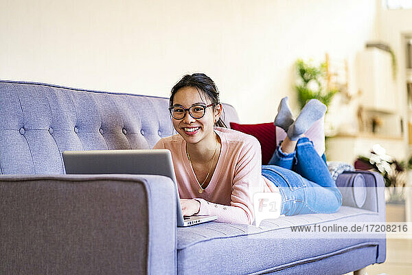 Lächelnde Frau liegt mit Laptop auf dem Sofa zu Hause