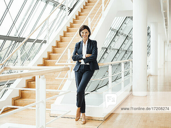 Smiling confident businesswoman with arms crossed by railing in corridor