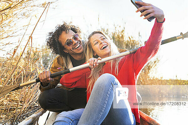 Glückliches Paar  das ein Selfie mit einem Ruder durch ein Mobiltelefon macht  während es in einem Kanu auf einem Fluss sitzt