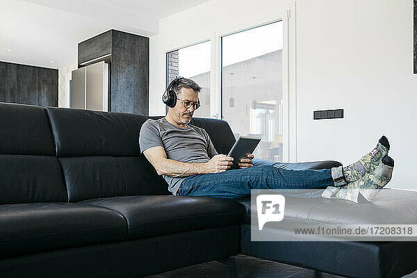 Älterer Mann hört Musik über Kopfhörer  während er ein digitales Tablet im Wohnzimmer benutzt