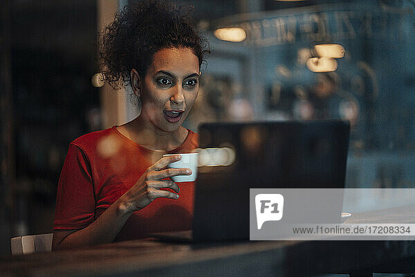Junge Frau hält eine Kaffeetasse und starrt mit offenem Mund auf einen Laptop in einem Café