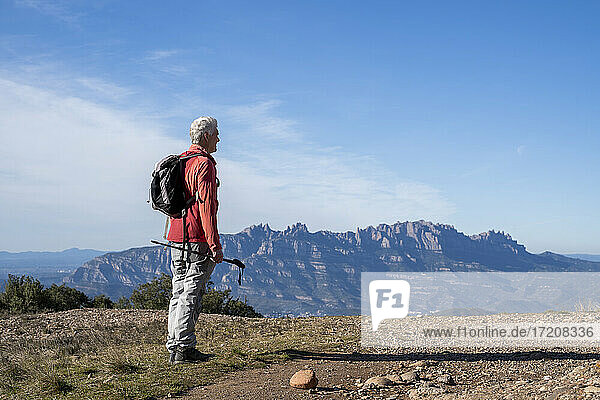 Älterer Mann mit Rucksack und Wanderstock mit Blick auf den Montserrat  während er auf einem Berg in Sant Llorenc del Munt i l'Obac  Katalonien  Spanien steht