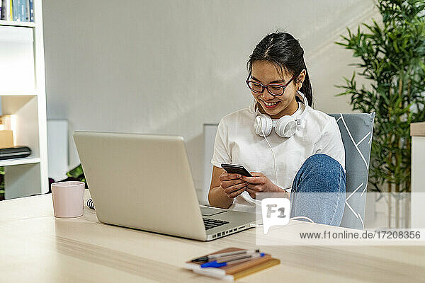 Junge Frau  die ein Mobiltelefon benutzt  während sie zu Hause am Laptop sitzt