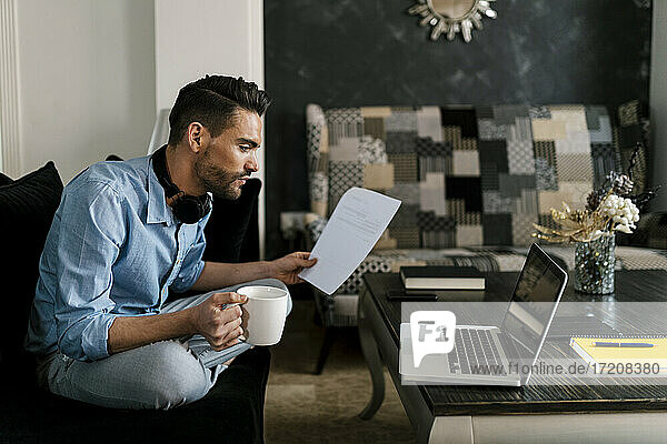 Geschäftsmann mit Kaffeetasse und Papier am Laptop  während er zu Hause sitzt