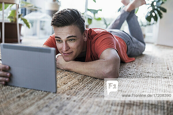 Glücklicher junger Mann mit digitalem Tablet auf dem Teppich liegend  während er im Wohnzimmer zu Hause ein Video ansieht