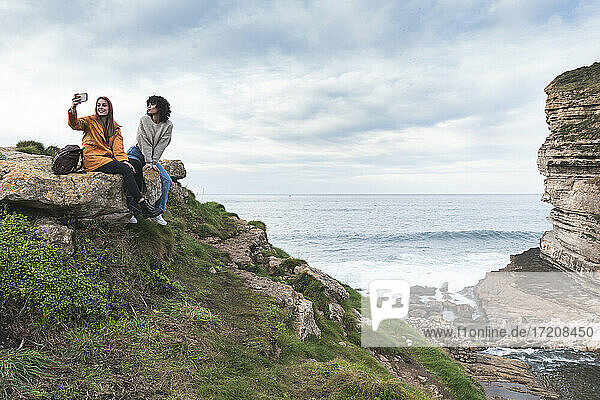Female friends taking selfie while sitting on cliff by sea at Acantilado El Bolao