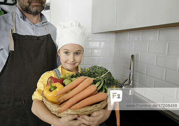 Lächelnde Tochter hält einen Gemüsekorb in der Hand  während sie bei ihrem Vater zu Hause steht