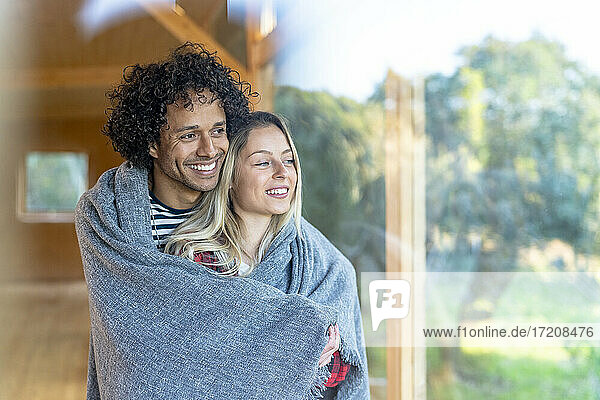 Lächelndes  in eine Decke gehülltes Paar  das zusammen am Fenster im Vorgarten steht