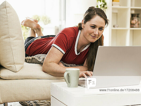 Frau benutzt Laptop  während sie zu Hause auf dem Sofa liegt
