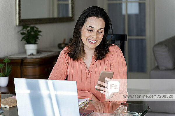Lächelnde Geschäftsfrau  die auf ihr Handy schaut  während sie am Esstisch im Heimbüro sitzt