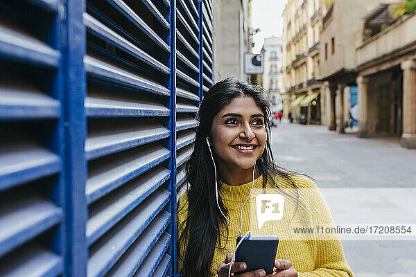 Lächelnde Frau mit Mobiltelefon  die wegschaut  während sie über Kopfhörer an einer blauen Wand Musik hört