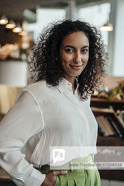 Junge Unternehmerin lächelt  während sie mit den Händen in den Taschen in einem Cafe steht