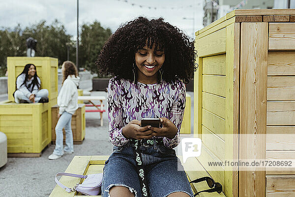 Smiling teenage girl using smart phone while sitting on box in city