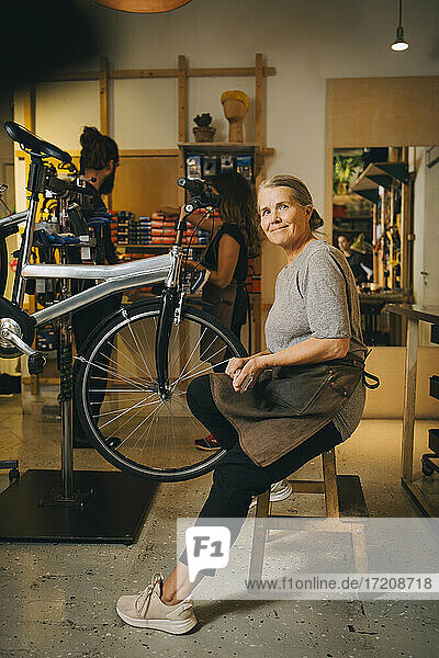Porträt eines lächelnden weiblichen Mechanikers  der auf einem Tisch in einem Fahrradgeschäft sitzt