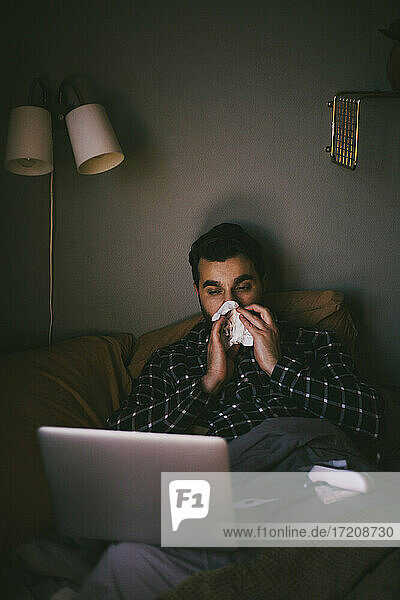 Kranker Mann schnäuzt sich die Nase  während er einen Laptop im Schlafzimmer zu Hause benutzt