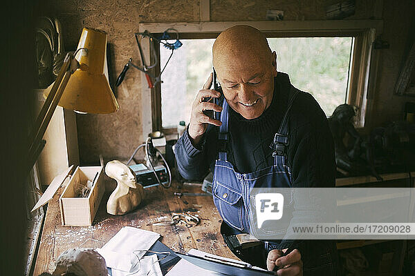 Lächelnder Geschäftsmann  der mit einem Smartphone in einer Werkstatt spricht