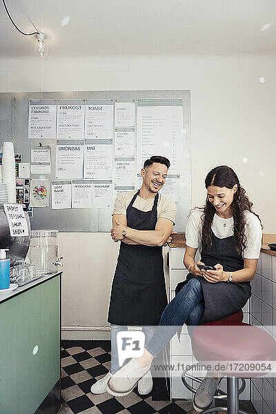 Female entrepreneur using smart phone while male colleague with arms crossed standing by in cafe