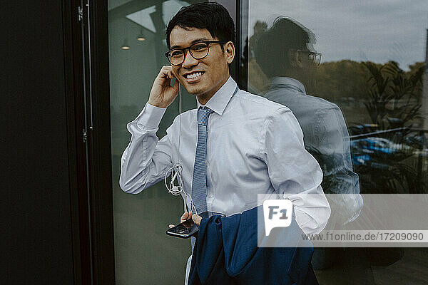 Porträt eines lächelnden Geschäftsmannes  der Musik über Kopfhörer hört  während er am Fenster steht