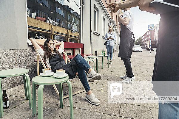 Lächelnde weibliche Besitzerin mit Händen hinter dem Kopf sitzt auf Liegestuhl außerhalb Kaffee-Shop