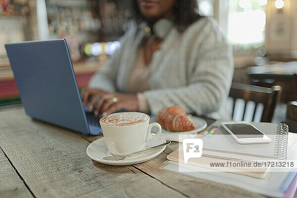 Frau arbeitet am Laptop neben Croissant und Cappuccino im Café