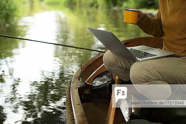 Mann Fliegenfischen und mit Laptop mit Kaffee im Boot auf dem Fluss
