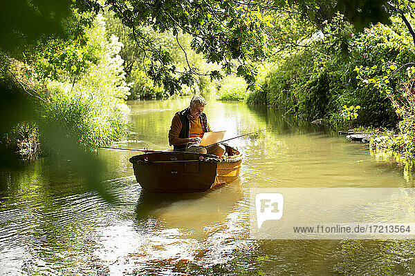 Man using laptop and fly fishing in boat on sunny river