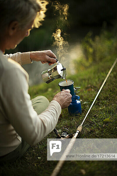 Man with fly fishing pole pouring coffee on riverbank