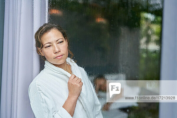 Neugierige Frau im Bademantel schaut aus regnerischem Fenster