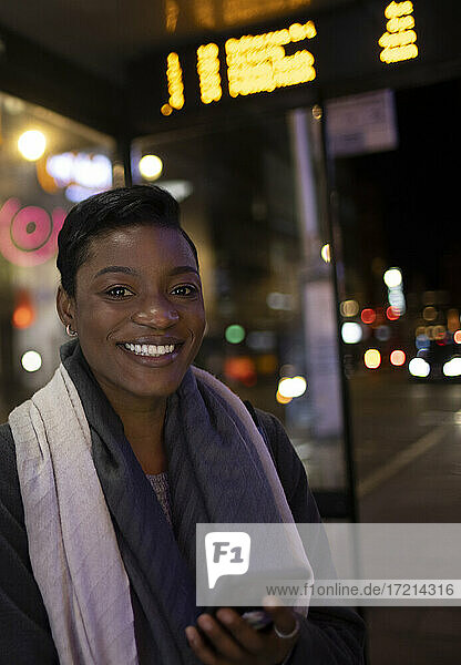 Portrait happy young woman with smart phone on city sidewalk at night