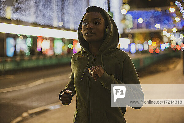 Young woman in hoody jogging on city street with lights at night